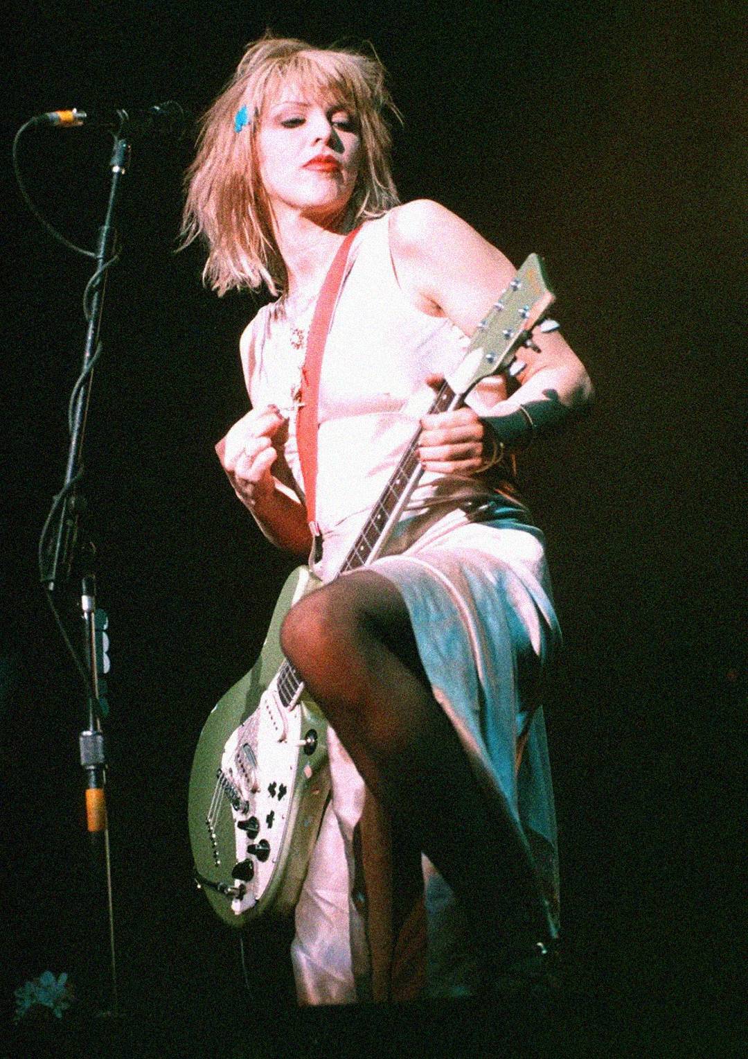 A woman with blond hair in a white dress plays an electric guitar on stage under dramatic lighting, standing in front of a microphone.