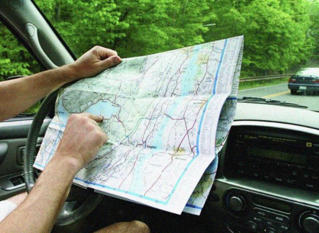 A person sitting in a car holds a large paper map over the steering wheel and points to a location. The view outside shows a road lined with trees and another car ahead.