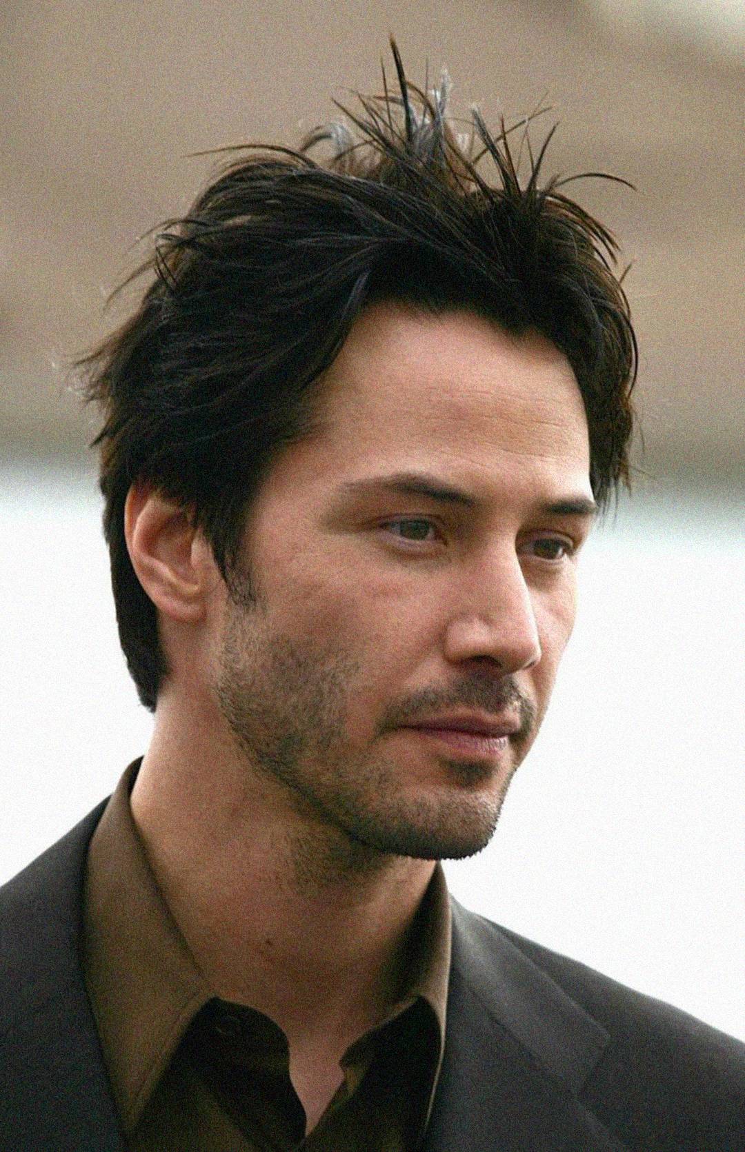 A man with tousled dark hair and light facial stubble wears a brown shirt and dark blazer, looking slightly downward with a thoughtful expression against a blurred, neutral background.