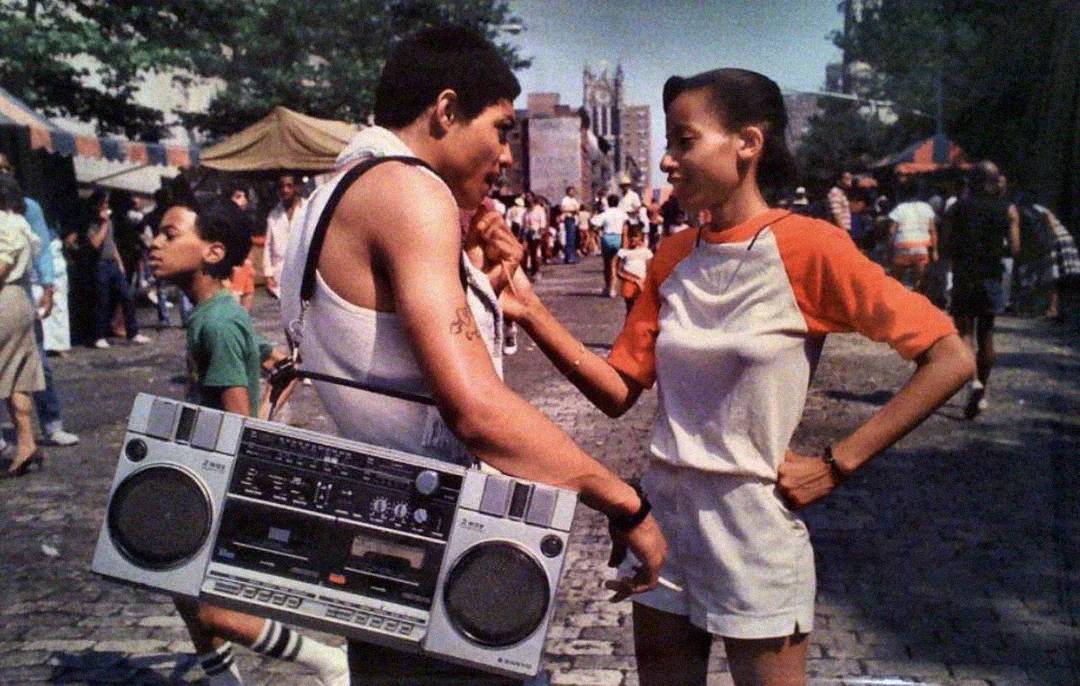Two young people talk on a cobblestone street at an outdoor event. One wears a tank top and carries a large boombox; the other wears a white and red T-shirt and shorts. People and tents are visible in the background.