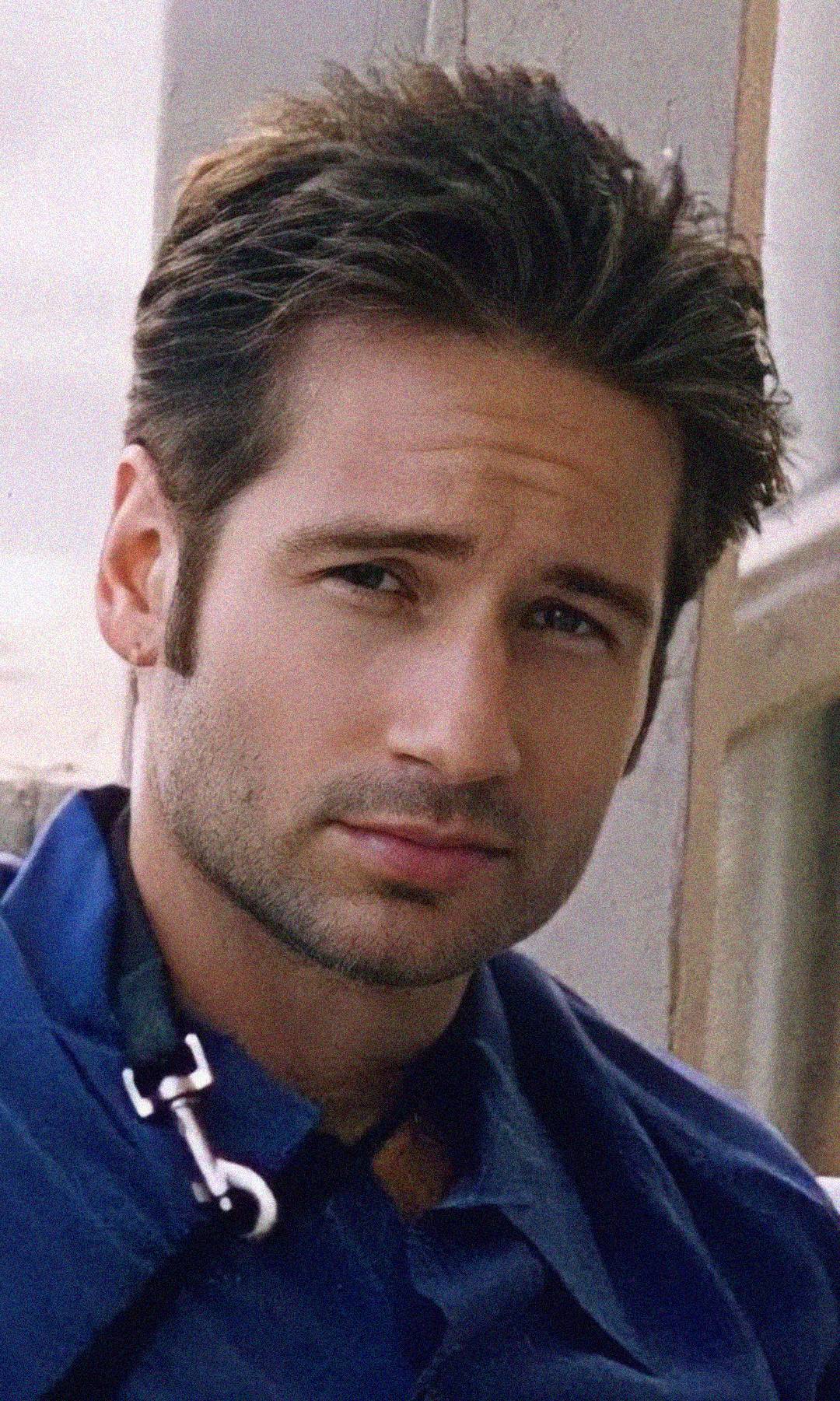 A man with short, dark hair and light facial stubble, wearing a blue jacket, looks directly at the camera with a slight, thoughtful expression.