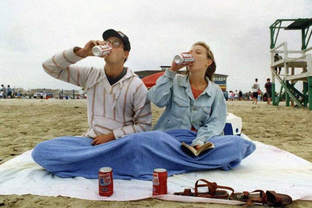 Two people sit on a blanket at the beach, covered by a blue blanket, drinking from soda cans. An open book, sandals, and soda cans are on the blanket. The beach is busy with people in the background.
