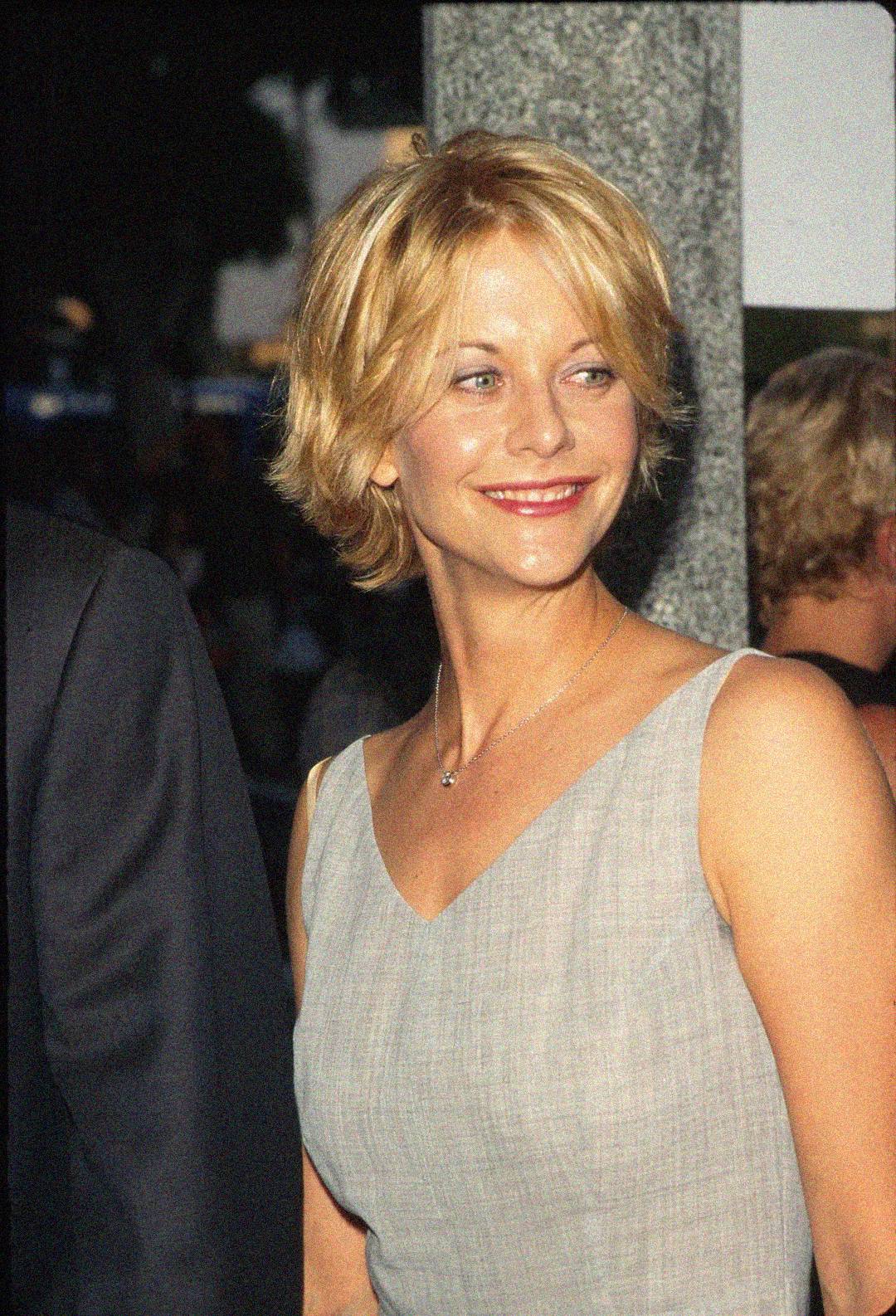 A woman with short blonde hair and a light gray sleeveless dress smiles while looking to the side at what appears to be a formal event.