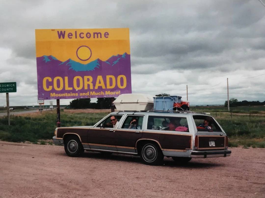 A vintage station wagon filled with luggage and people drives past a large "Welcome to Colorado: Mountains and Much More!" sign under a cloudy sky. The road is rural with open fields on either side.