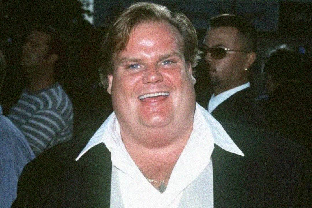 A smiling man with light skin and blond hair wears a white shirt with an open collar and a dark jacket. He is photographed outdoors, with people and blurred surroundings in the background.