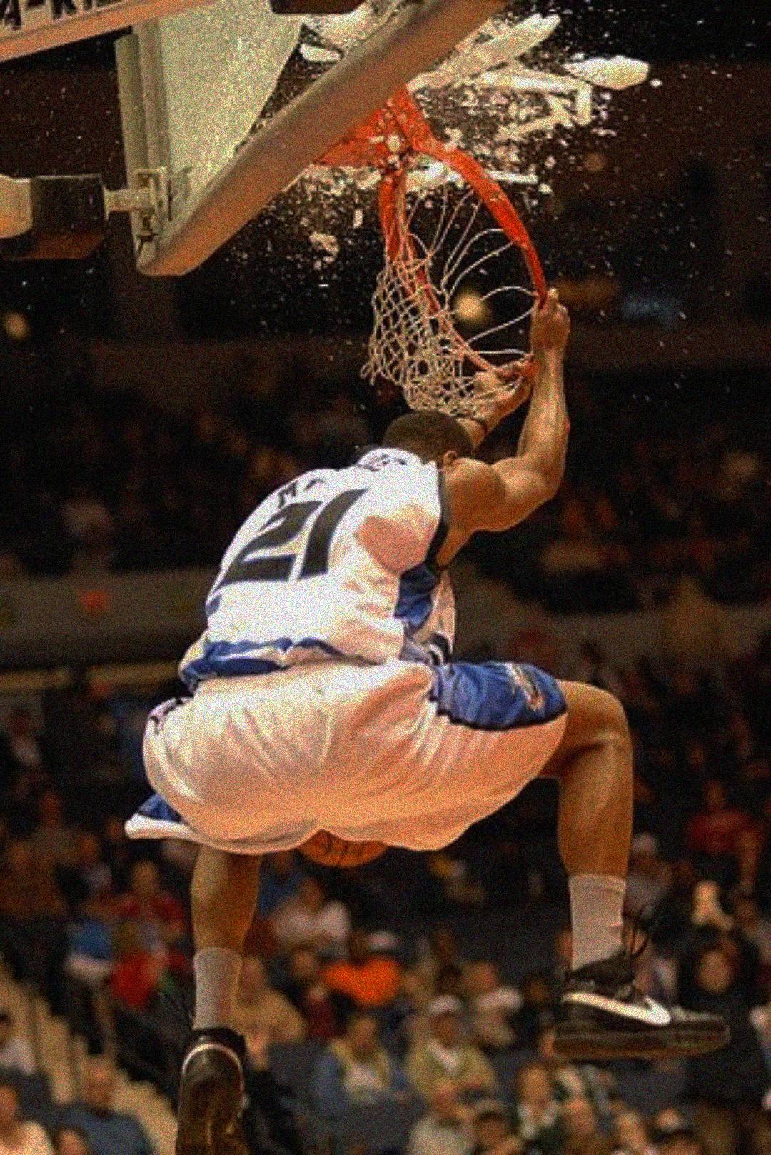 A basketball player in a white and blue uniform slams a dunk, breaking the backboard glass, with shards flying as the crowd watches in the background.