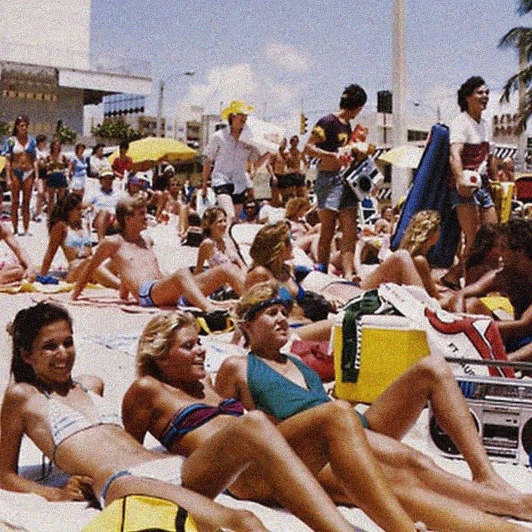 A crowded beach scene with many people sunbathing, lounging on towels, and relaxing under umbrellas on a sunny day. Most are in swimwear, enjoying the warm weather. Buildings and palm trees are visible in the background.