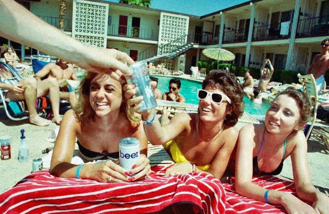 Three women in swimsuits are smiling and lounging by a pool, holding drinks, while a hand offers a can to one of them. People are relaxing and sunbathing in the background at a retro-style outdoor pool.