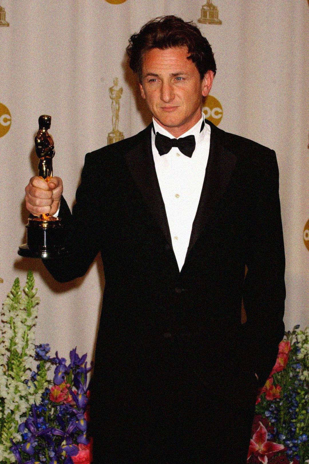 A man in a black tuxedo stands in front of a backdrop holding up an Oscar trophy, surrounded by flowers at the bottom of the image.