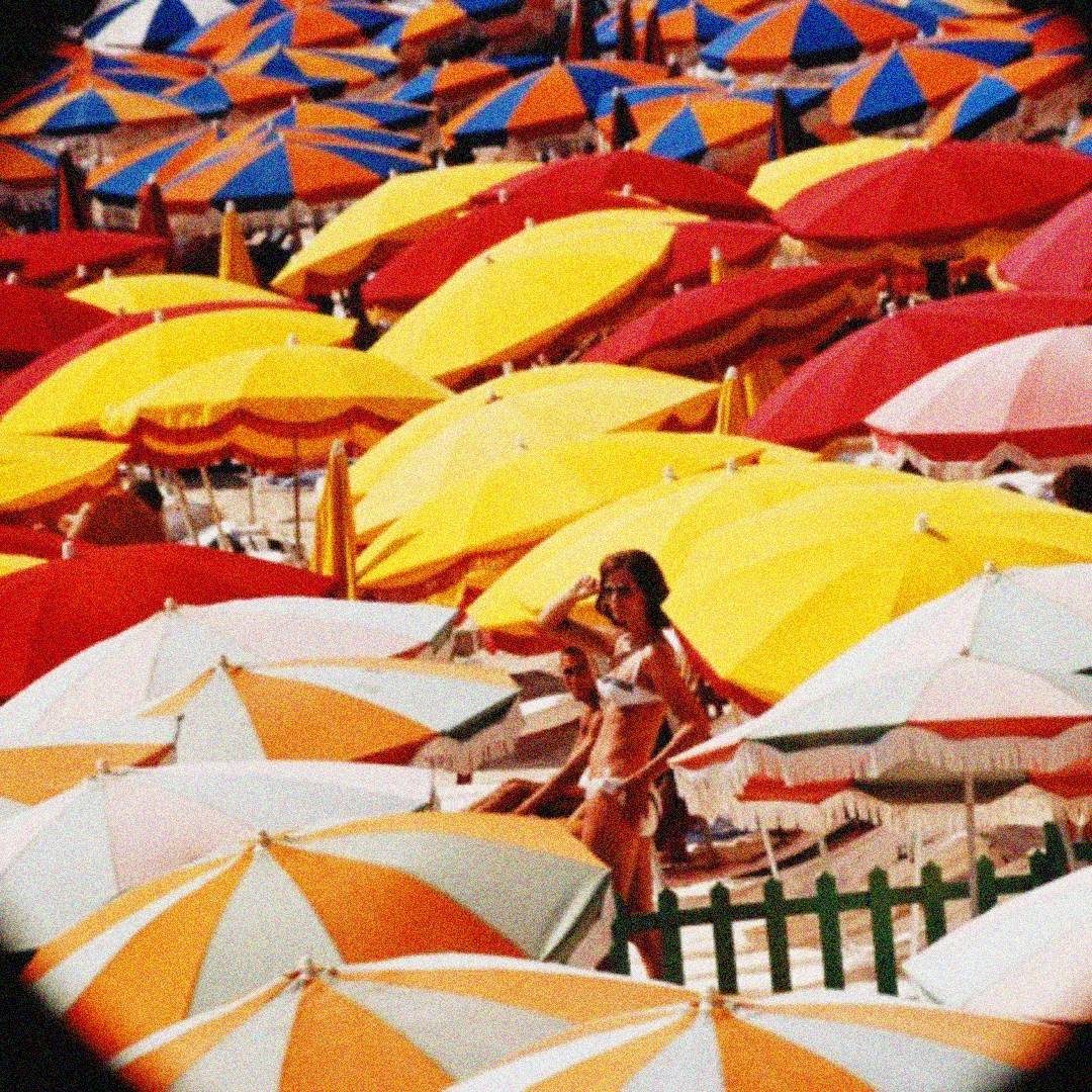 A woman in a white bikini stands among a crowded beach filled with colorful umbrellas in yellow, orange, and white. The umbrellas cover most of the sand, creating a vibrant, summery scene.