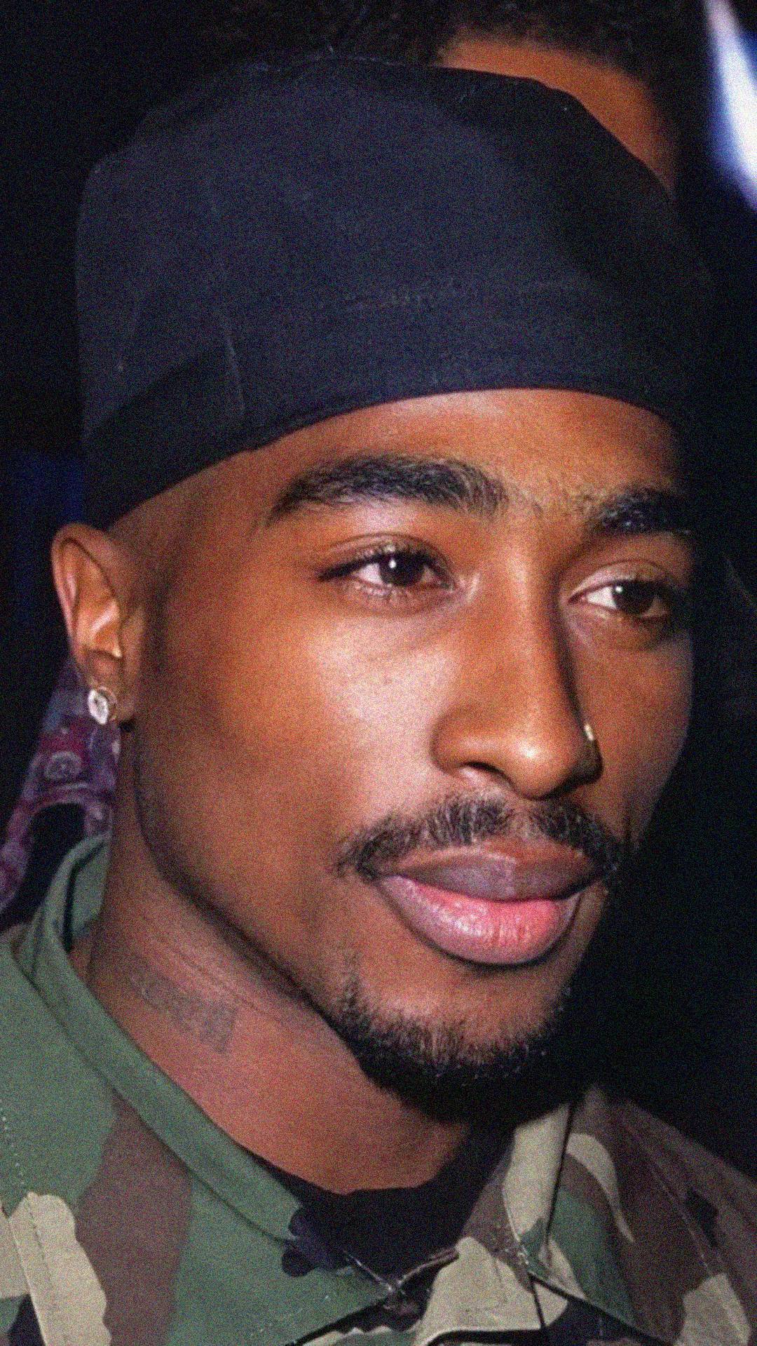 A man with a thin mustache and beard wears a black bandana, diamond earring, nose ring, and camouflage clothing, looking slightly to the side with a neutral expression.