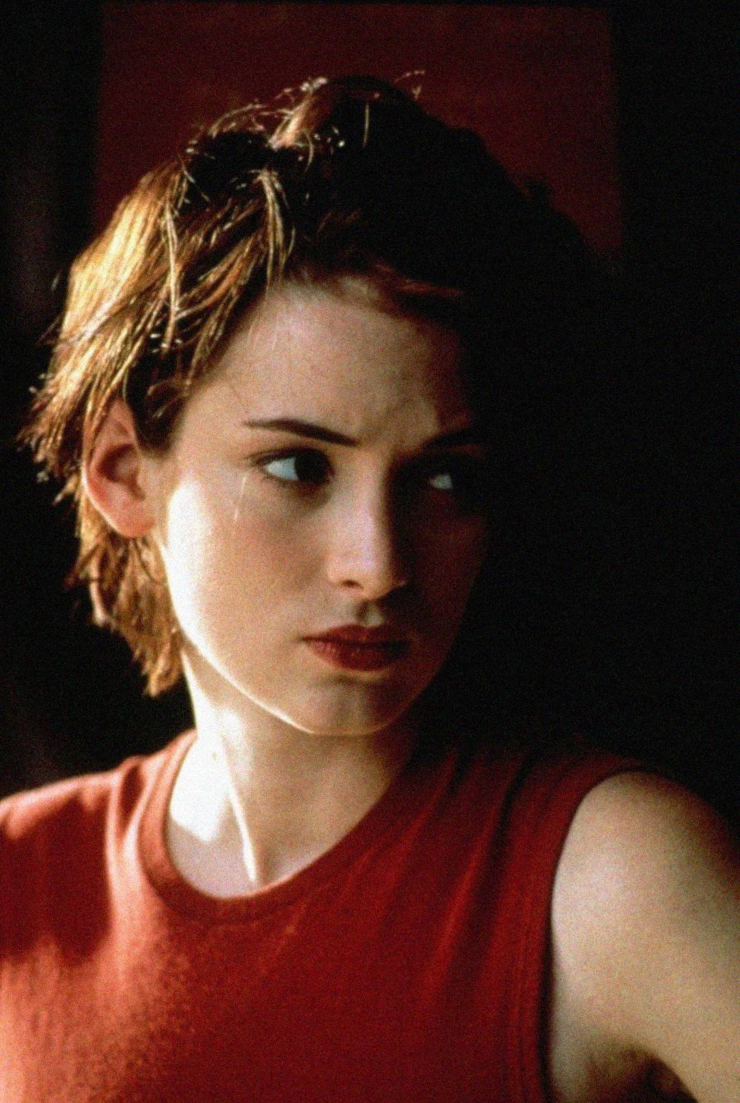 A young woman with short brown hair wearing a sleeveless red top looks to the side with a serious expression, against a dark background.