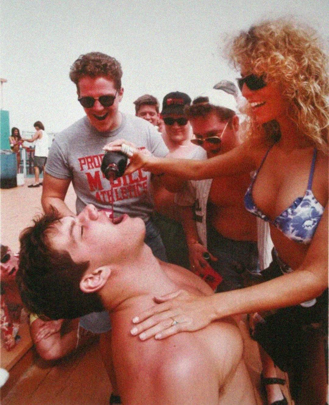 A woman in a blue bikini pours a drink into a shirtless man's mouth as several people in sunglasses and casual clothes smile and watch on a sunny outdoor deck.