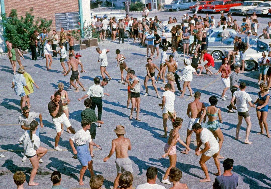 A large group of people in swimsuits and summer clothing dance and mingle outdoors in a parking lot, with classic cars and spectators in the background on a sunny day.