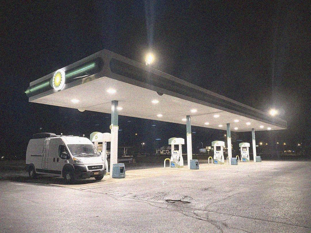 A white van is parked at a nearly empty gas station at night, illuminated by bright overhead lights. Four gas pumps are visible under the canopy, and the surrounding area appears quiet and deserted.