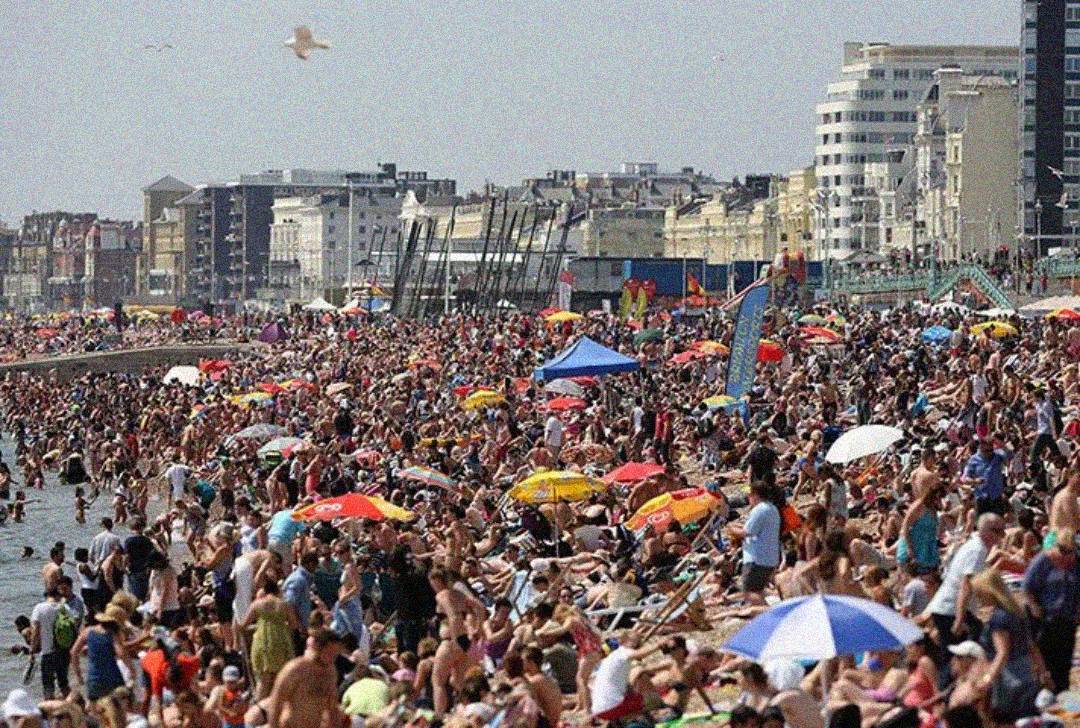 A crowded beach scene with thousands of people sunbathing, swimming, and sitting under colorful umbrellas. Buildings and apartments line the background under a bright sky.