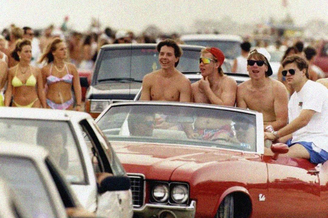 Four young men in swimwear ride in a red convertible with the top down, smiling and enjoying a crowded beach scene. Other people in swimsuits walk and stand near cars in the background.