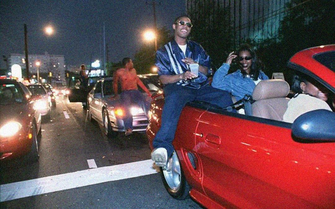 Two people sit on the back of a red convertible stopped in traffic at night; one holds a drink, both wear sunglasses and appear relaxed, while cars line the street behind them under city lights.