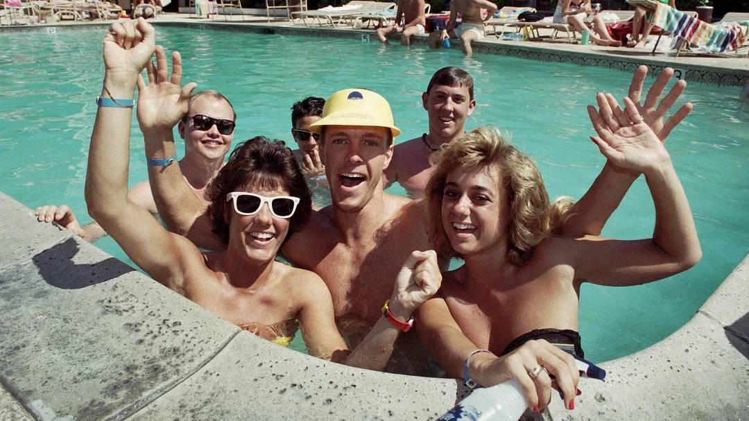A group of smiling people in swimsuits wave at the camera while standing together in a swimming pool, with lounge chairs and more people in the background.
