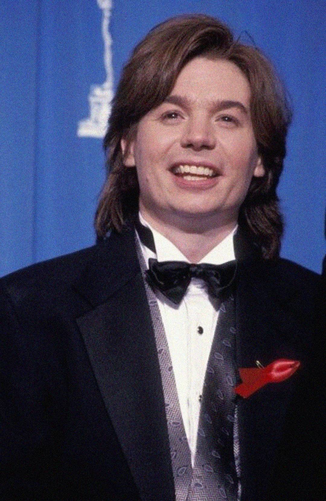A person with medium-length brown hair wearing a black tuxedo, bow tie, and a shiny vest smiles at the camera. A red ribbon is pinned to the jacket. The background is blue with a white outline of a statue.