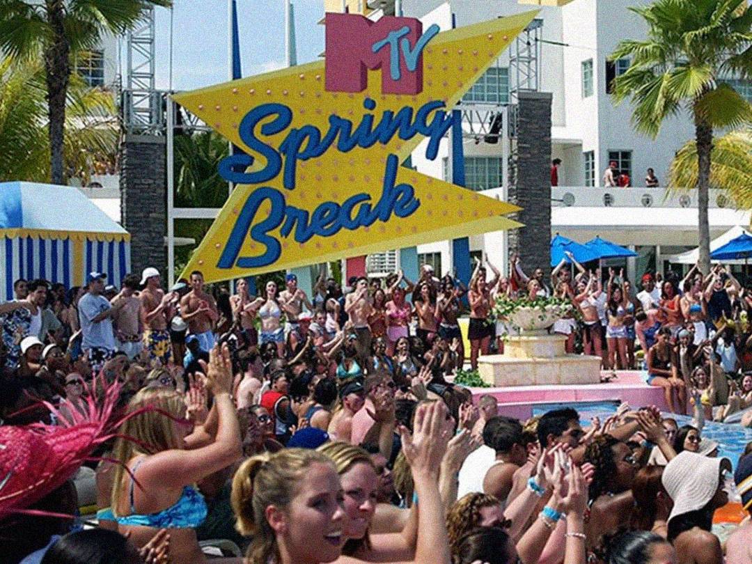 A large crowd of people in swimsuits cheer and clap around a pool under palm trees, with a colorful “MTV Spring Break” sign displayed in the background. The atmosphere is lively and festive.