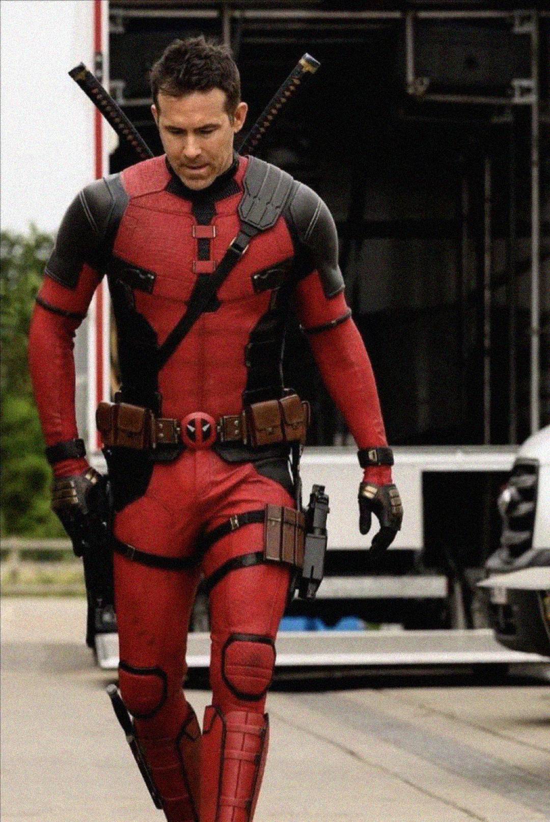 A person in a red and black superhero costume with swords on their back walks outdoors near trailers and vehicles.