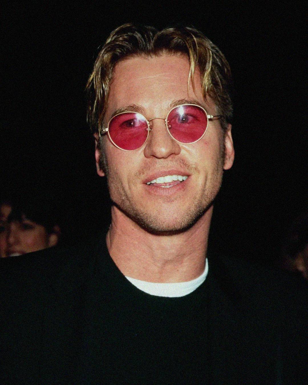 A man with blond hair wearing round pink sunglasses and a black jacket over a white shirt smiles in front of a dark background.