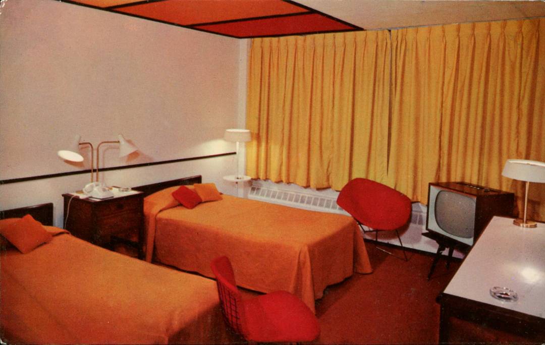 A retro hotel room with two twin beds, orange bedding, yellow curtains, red chairs, a vintage TV on a stand, and modern lamps, all in a coordinated orange and yellow color scheme.