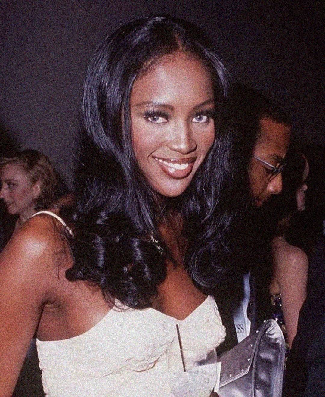 A woman with long, wavy black hair and a white dress smiles at the camera while holding a drink. She is in a crowded indoor setting with people in the background.