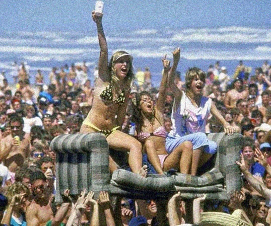 Three young women in swimsuits celebrate on a couch held above a crowded beach, surrounded by people in swimwear. The ocean waves are visible in the background.