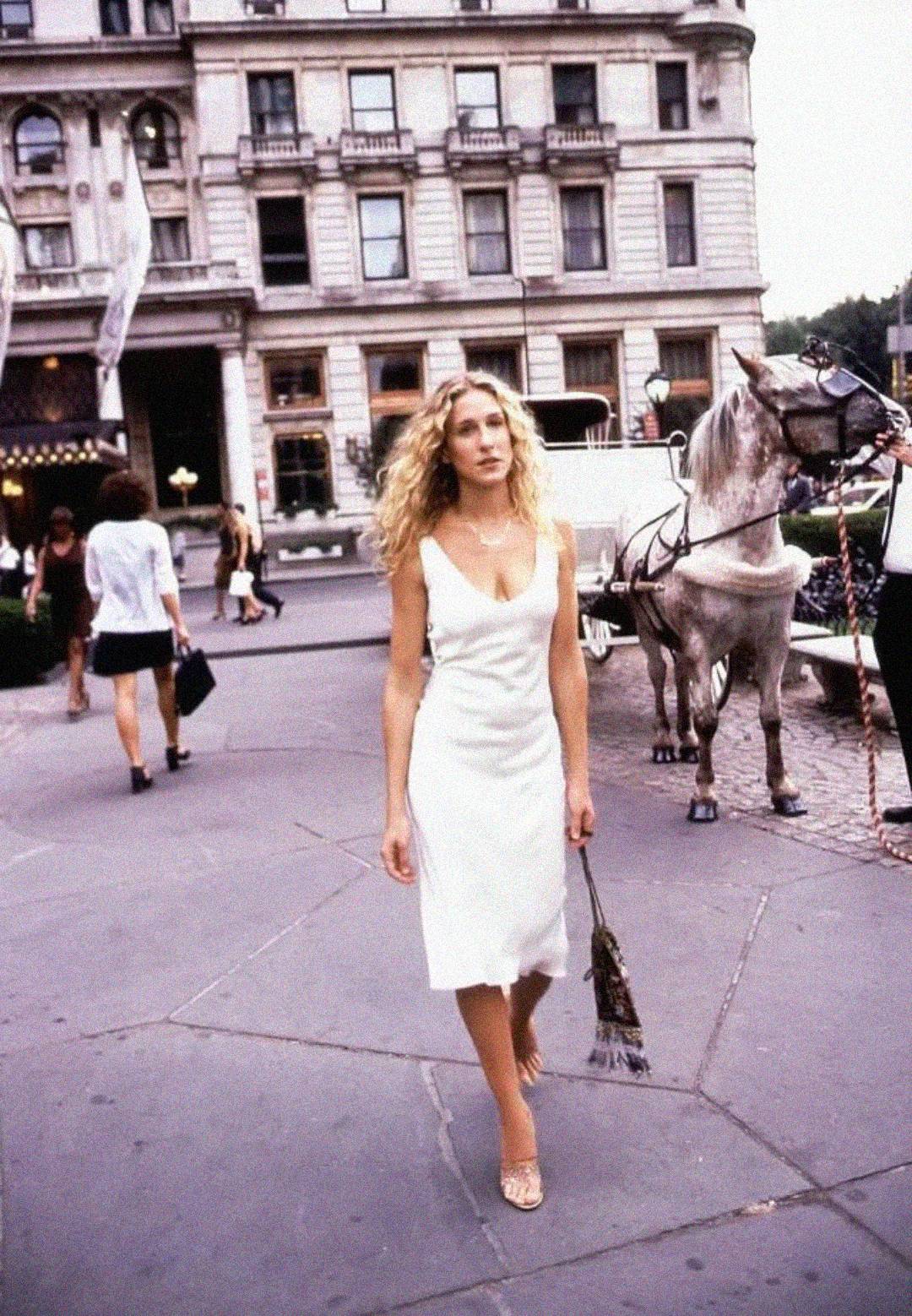 A woman with curly blonde hair in a white dress walks on a city sidewalk near a horse and carriage, with tall buildings and people in the background.