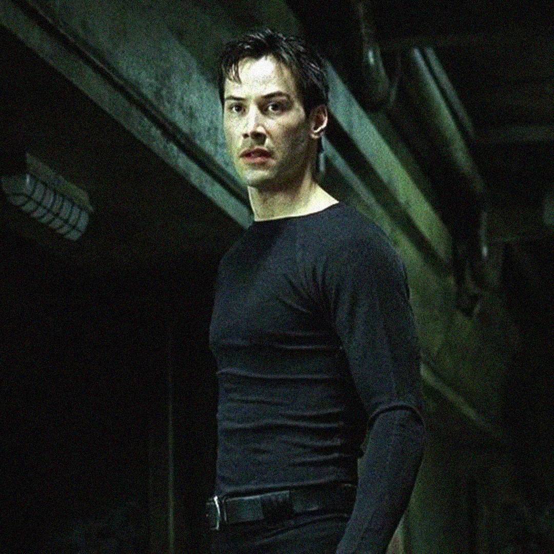 A man with short dark hair stands in a dimly lit, industrial-looking hallway. He wears a fitted black long-sleeve shirt and black pants, and looks ahead with a serious expression.
