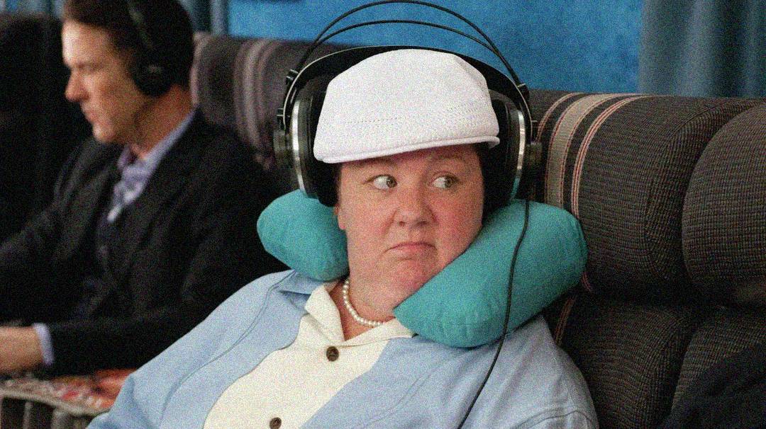 A woman wearing a white cap, headphones, and a neck pillow sits on an airplane, looking to the side with a skeptical expression. The seat beside her is occupied by a man wearing headphones.
