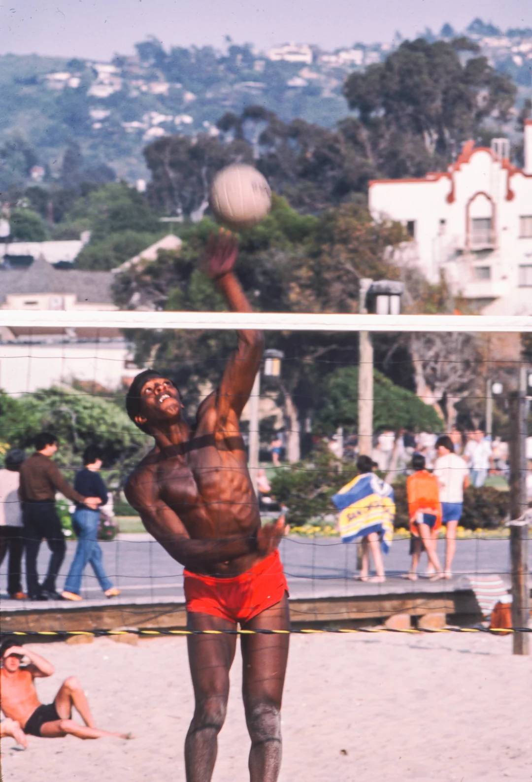 A man in red shorts jumps to hit a volleyball on a sandy outdoor court, with people and buildings in the background on a sunny day.
