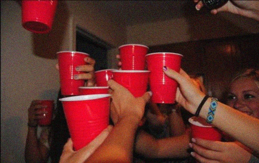 A group of people raising red plastic cups in a toast at a lively indoor gathering, with hands overlapping and a festive atmosphere.