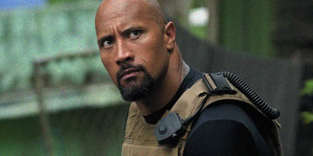 A muscular man with a bald head and goatee, wearing a tactical vest with a radio earpiece, looks intensely to the side in an outdoor setting with blurred greenery in the background.