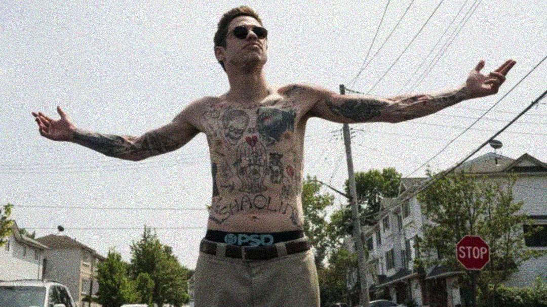 A shirtless man with tattoos on his chest and arms stands in the middle of a street with arms outstretched, wearing sunglasses and khaki pants. Houses, trees, power lines, and a stop sign are visible in the background.