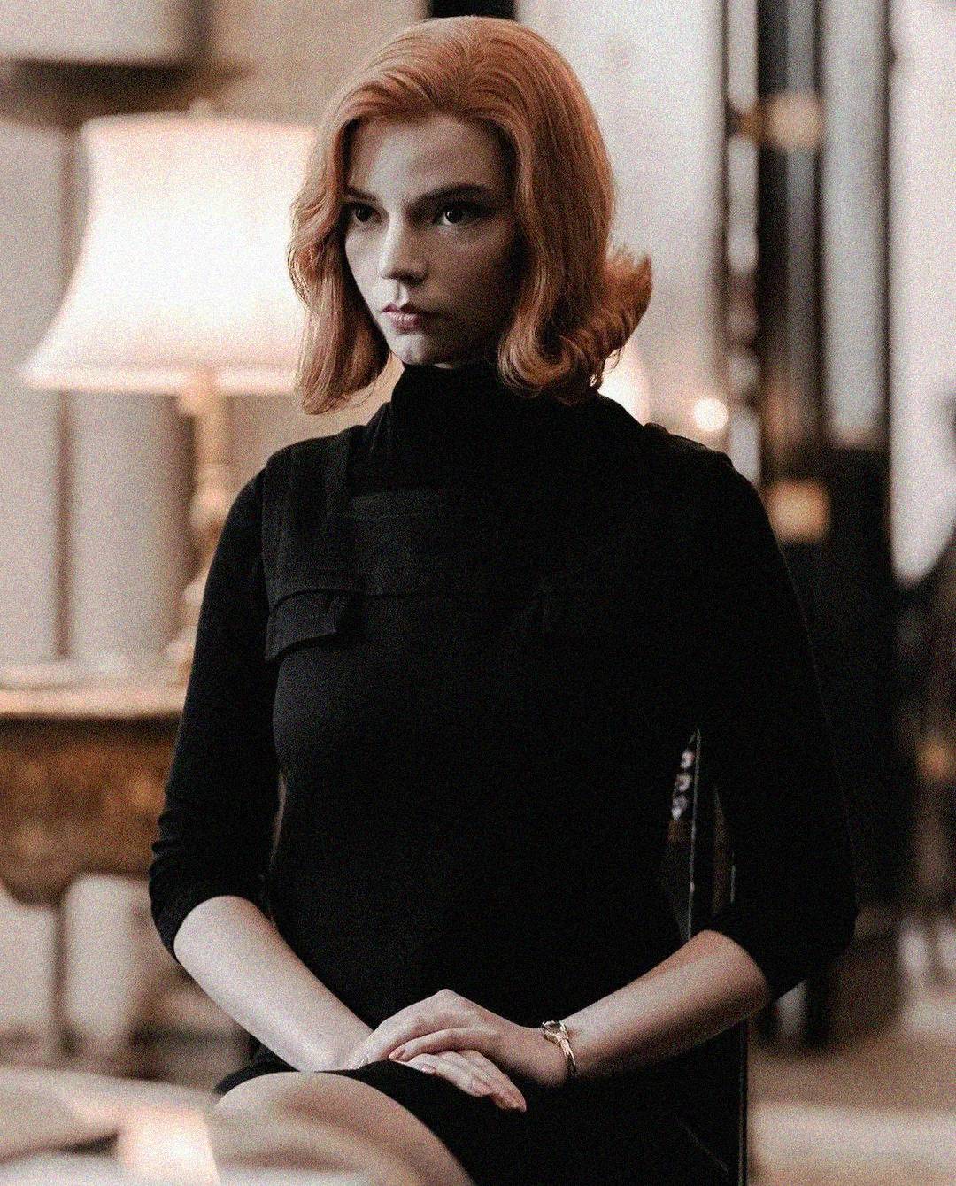 A woman with shoulder-length red hair, wearing a black dress, sits with her hands folded on her lap in a softly lit room with a table lamp and wooden furniture in the background.