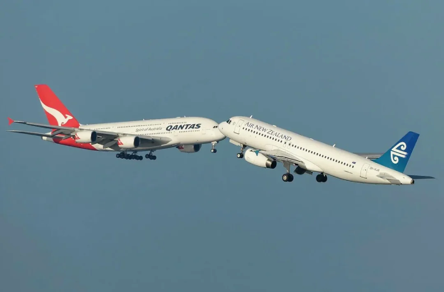 A Qantas airplane and an Air New Zealand airplane appear to be flying extremely close to each other in the sky, almost touching, against a clear blue background.