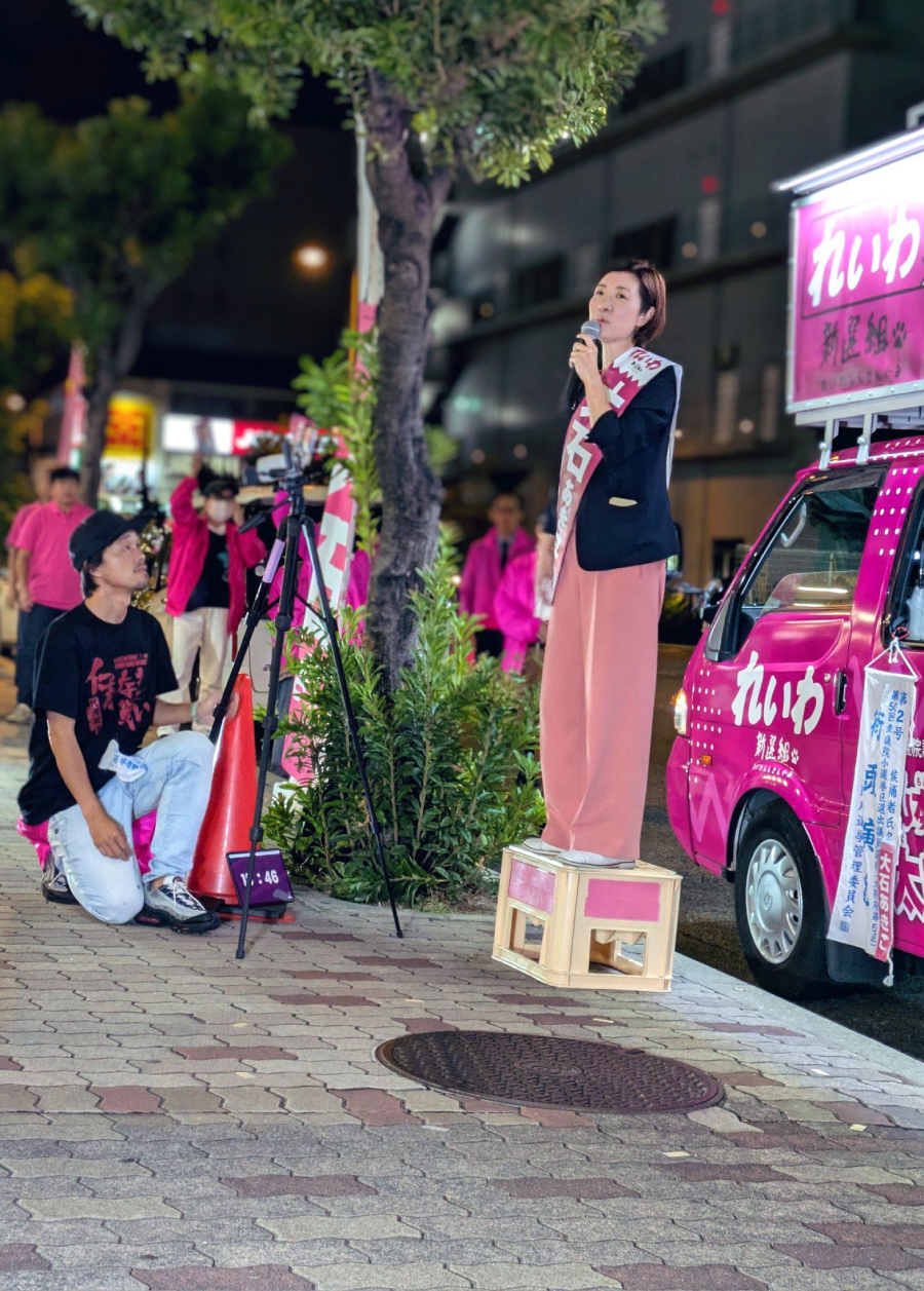 A woman stands on a box, speaking into a microphone during an outdoor nighttime event. She wears a sash and is next to a pink van with Japanese writing. Several people in pink clothing stand nearby. A cameraman kneels, filming her.
