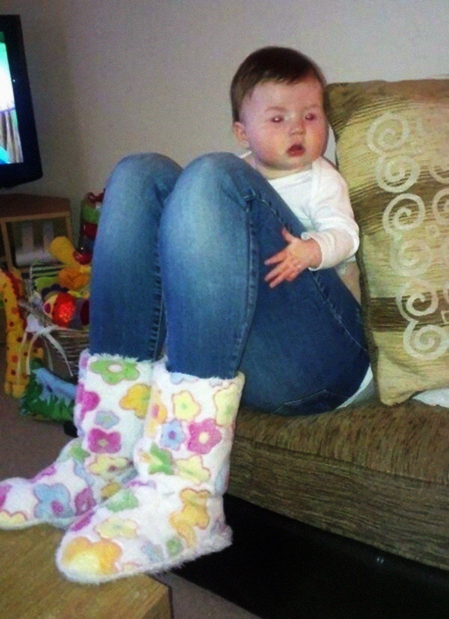 A baby’s head has been humorously edited onto an adult’s body wearing jeans and colorful fluffy slippers, sitting on a couch with knees pulled up, creating a funny and surreal effect.