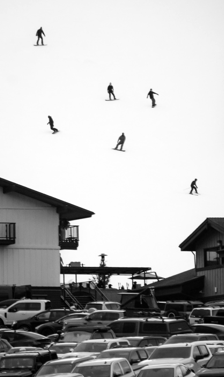 Black and white image of six snowboarders or skiers descending a snowy slope above buildings and a parking lot filled with cars, with the scene viewed from a distance.
