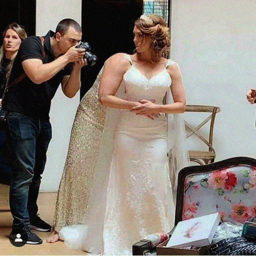 A bride in a white gown stands indoors, hands clasped, as a photographer crouches nearby taking photos. Several people are around her and floral and decorative items are visible in the foreground.