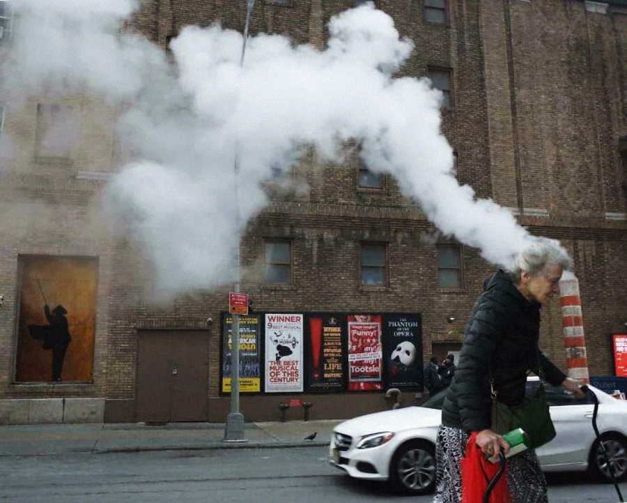 An elderly woman walks past a street steam vent, making it appear as if white steam is coming out of her head. Brick building and colorful posters are visible in the background.