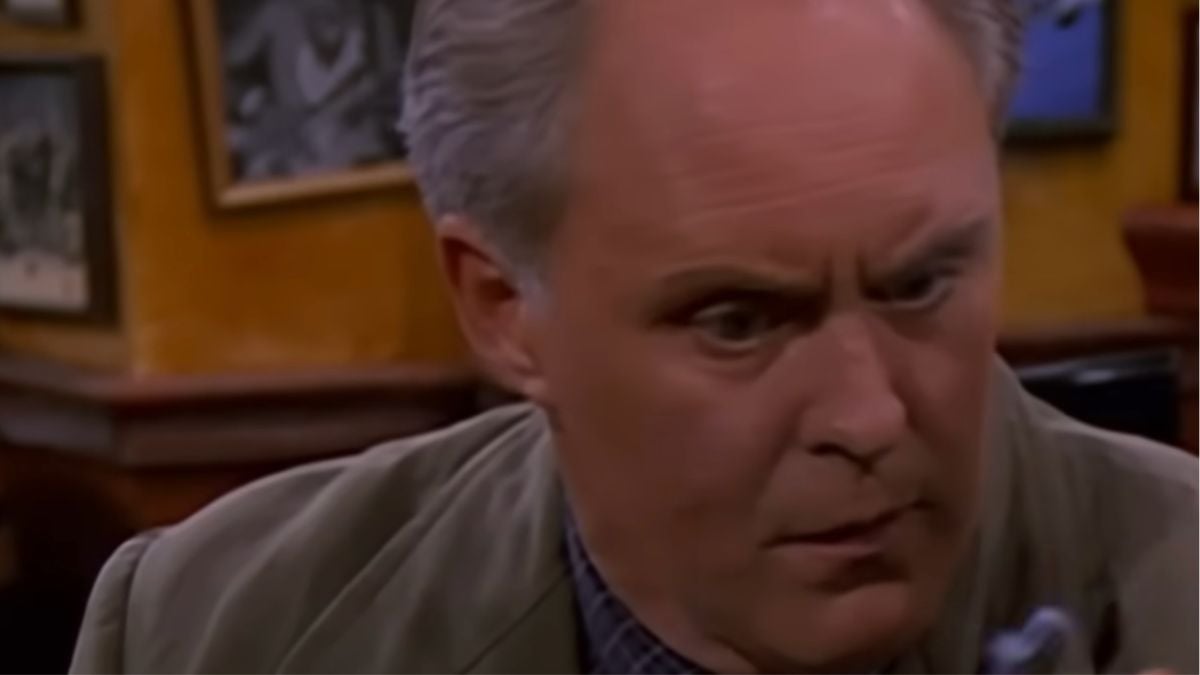 A man with gray hair looks down with a surprised and intense expression in a room with framed pictures on the yellow walls behind him.