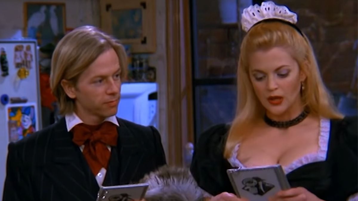 A man in a bow tie and suit stands next to a woman dressed as a maid with a white headpiece. Both are holding menus and appear to be talking in a kitchen setting.