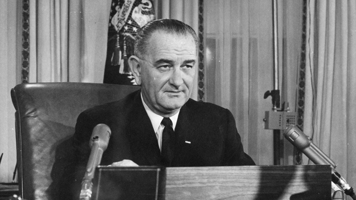 A man in a suit sits at a desk with microphones in front of him, appearing to give a speech or address. Curtains and part of a flag are visible in the background.