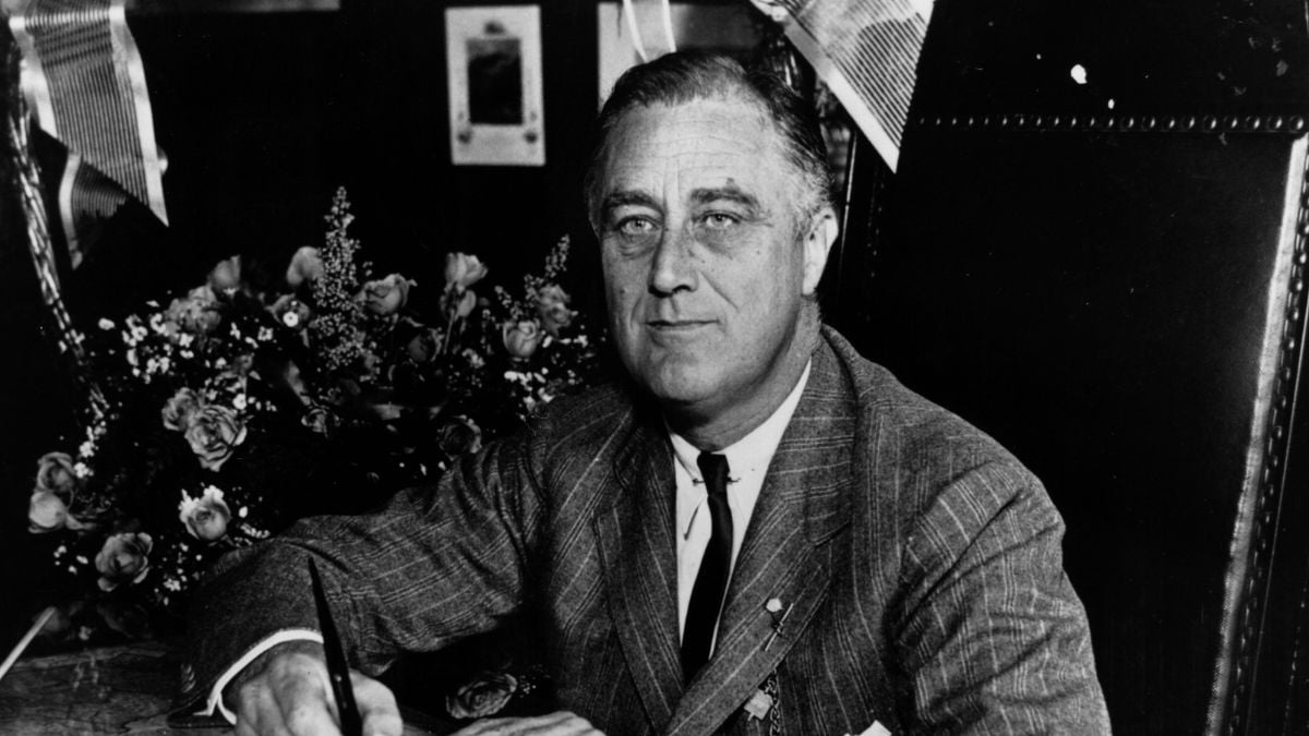 A man in a suit and tie sits at a desk, holding a pen, with flowers and papers in the background. He looks directly at the camera with a neutral expression.