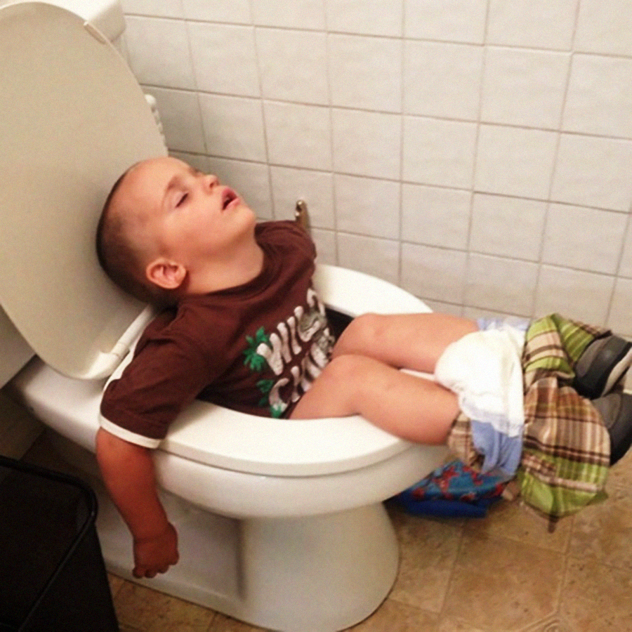 A young boy, wearing a brown shirt and plaid shorts, is asleep sitting on a toilet with his head resting against the seat and his pants pulled down, appearing to have dozed off while using the bathroom.