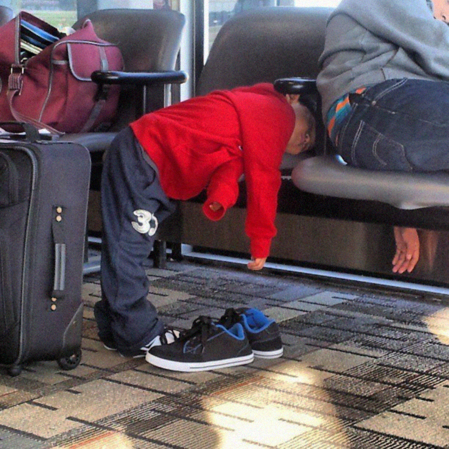 A young child in a red shirt leans and rests his head on an airport bench while standing, with shoes off and suitcase nearby, next to an adult seated on the bench.